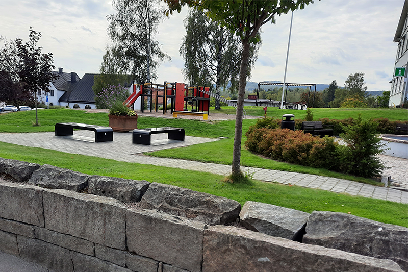 På bilden visas parken med en del av en fontän och parkbänkar som möjliggör mobilladdning med hjälp av solceller. I bakgrunden syns en större klätterställning. 