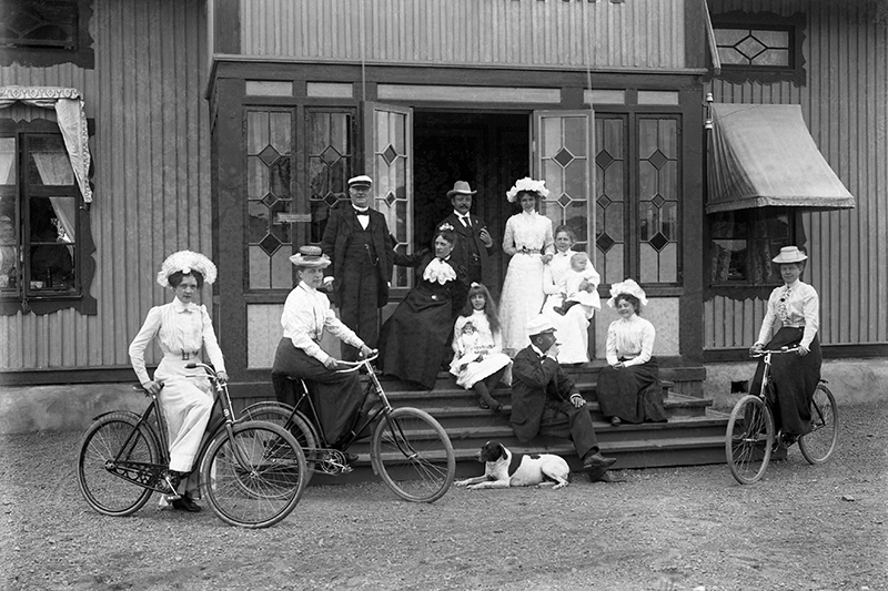 En familj i gamla kläder framför en vill, två kvinnor sitter på cykel.